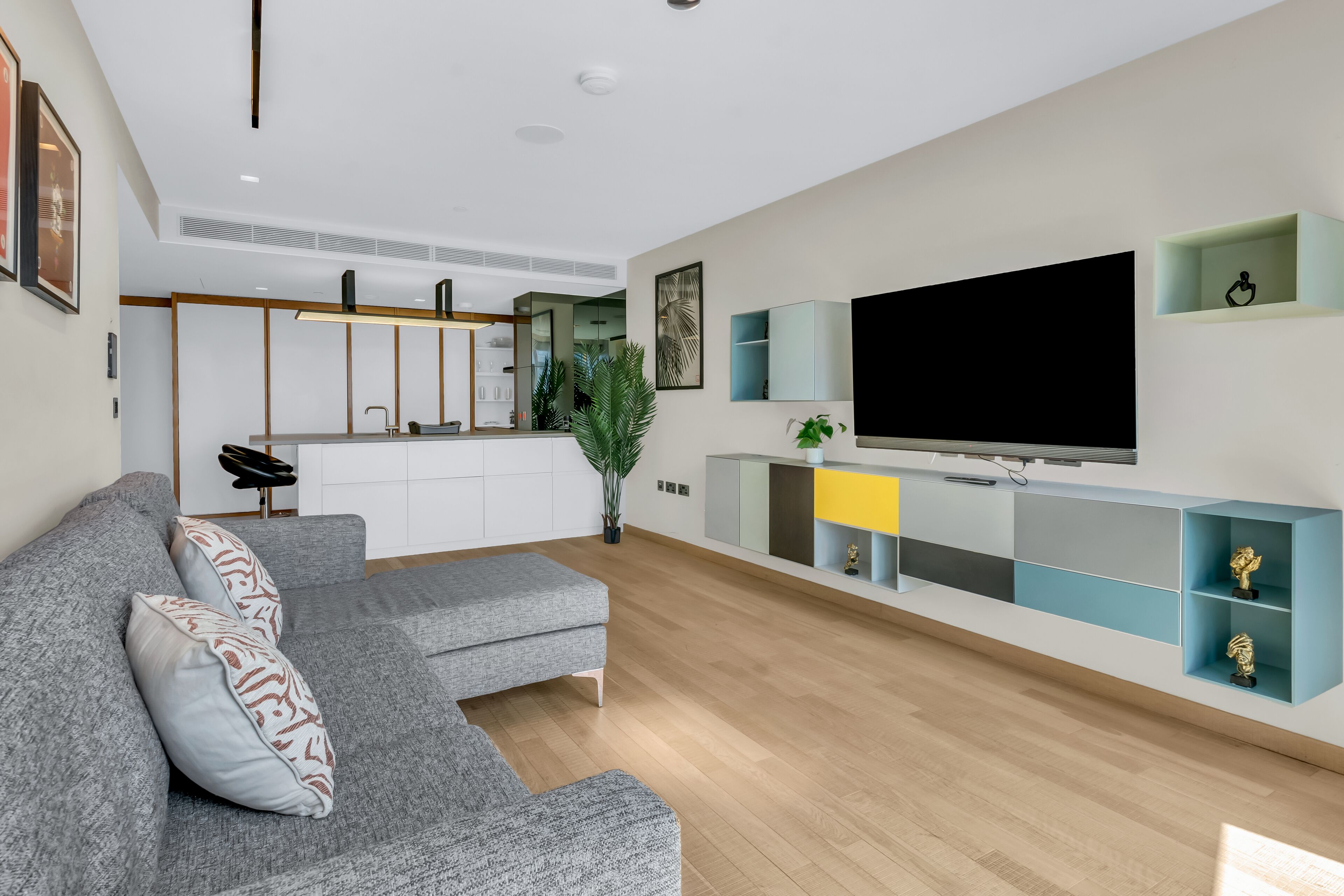 A modern living room with a grey sectional sofa, a flat-screen TV mounted on a wall, and a minimalist kitchen in the background.