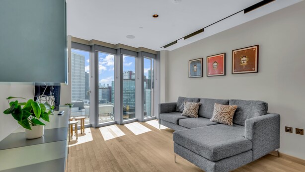 Una sala de estar moderna con un sofá gris, una mesa de centro de vidrio y una planta en maceta. Hay ventanas amplias que ofrecen vistas de la ciudad.
