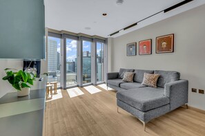 A modern living room with a grey sofa, a glass coffee table, and a potted plant. There are large windows offering a view of the cityscape. - Modern 1 Bedroom Apartment in Stratford (London)