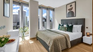 Un dormitorio moderno con una cama grande, una mesita de noche, una ventana con vista a la ciudad y un florero con flores.