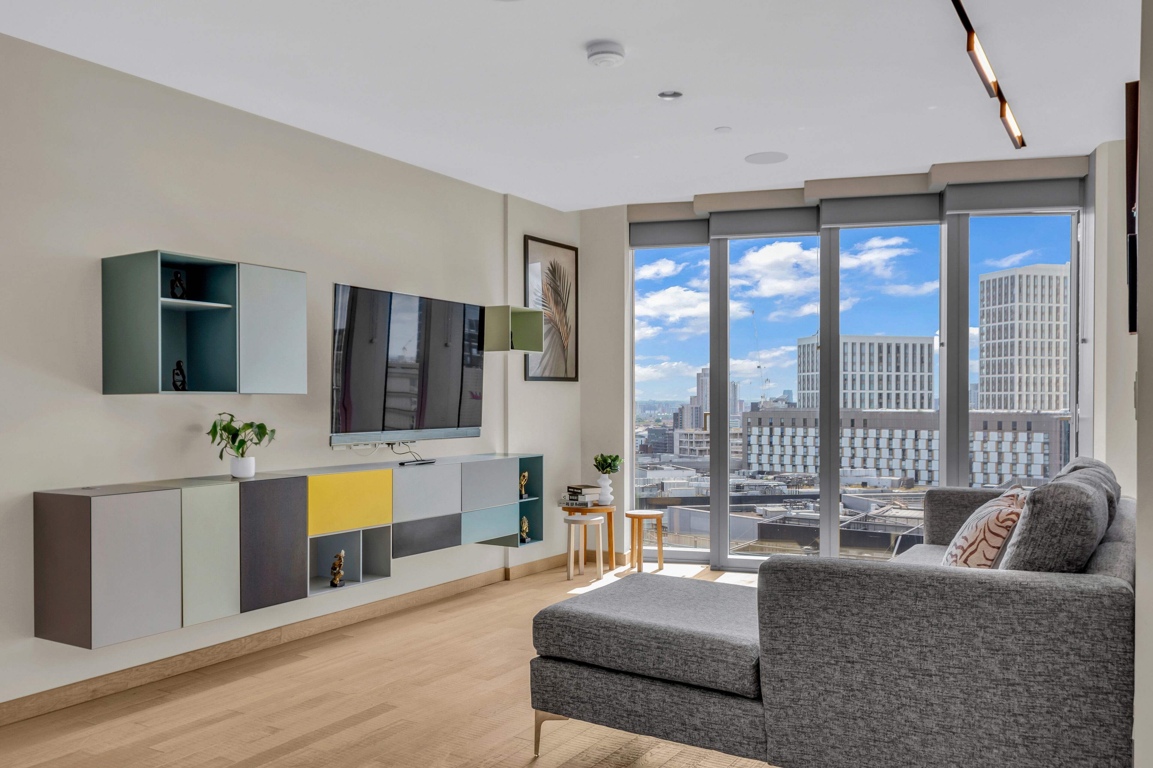 A modern living room with a grey sofa, a TV mounted on the wall, and a large window offering a city view.