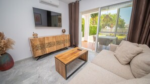 A living room with a sofa, a wooden coffee table, a TV mounted on the wall, a cabinet with intricate patterns, and a sliding glass door leading to an outdoor area with a table and chairs. - Los Pelicanos apt 2.103 (Manilva)