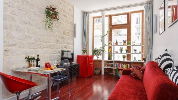 Ein gemütliches Wohnzimmer mit einem roten Sofa, einem kleinen Esstisch mit Stühlen, einem roten Kühlschrank und einem Bücherregal voller Bücher und Pflanzen.