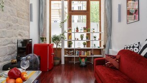 Ein gemütliches Wohnzimmer mit einem roten Sofa, einem kleinen roten Kühlschrank, einem Holztisch mit Obst und einer Bücherwand voller Bücher und Pflanzen.