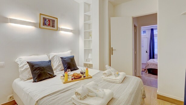Une chambre à coucher avec un lit, des oreillers, un plateau contenant des articles pour le petit-déjeuner, une table de chevet avec une lampe et une porte donnant sur une autre pièce.
