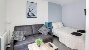 Quarto moderno e compacto com sofá cinza, cama branca, uma mesinha com uma planta e uma obra de arte emoldurada na parede.