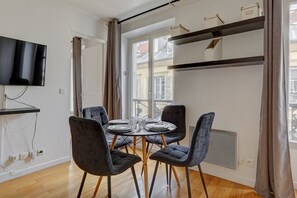 Un coin repas avec une table dressée pour quatre personnes, quatre chaises grises à revêtement rembourré, un téléviseur à écran plat fixé au mur et une fenêtre avec des rideaux.