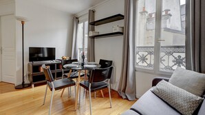 A modern living room with a dining area, a TV, and a balcony with decorative railings. - 395 Suite BIOT 10 - Superb apartment in Paris (Paris 17 Batignolles-Monceau)