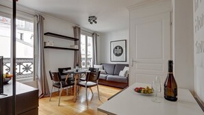 A modern living room with a dining area, a sofa, a table with a bottle and glasses, and a balcony with a railing. - 395 Suite BIOT 10 - Superb apartment in Paris (Paris 17 Batignolles-Monceau)