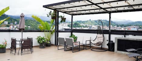 A rooftop terrace with a covered seating area, potted plants, and a view of a cityscape and mountains.