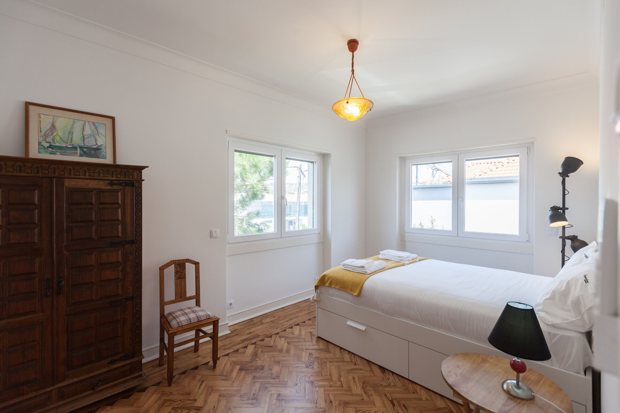Een slaapkamer met een bed, een houten garderobe, een stoel, een nachtkastje met een lamp en een raam.