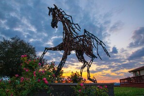 A metal sculpture of a horse in a garden with blooming flowers and a sunset in the background.