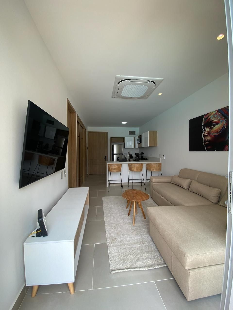 A modern living room with a flat-screen TV, a white console table, a beige sofa, and a small wooden coffee table. A kitchen with a dining area is visible in the background.