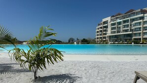 A beach with a clear blue pool, palm trees, and a multi-story hotel building in the background.