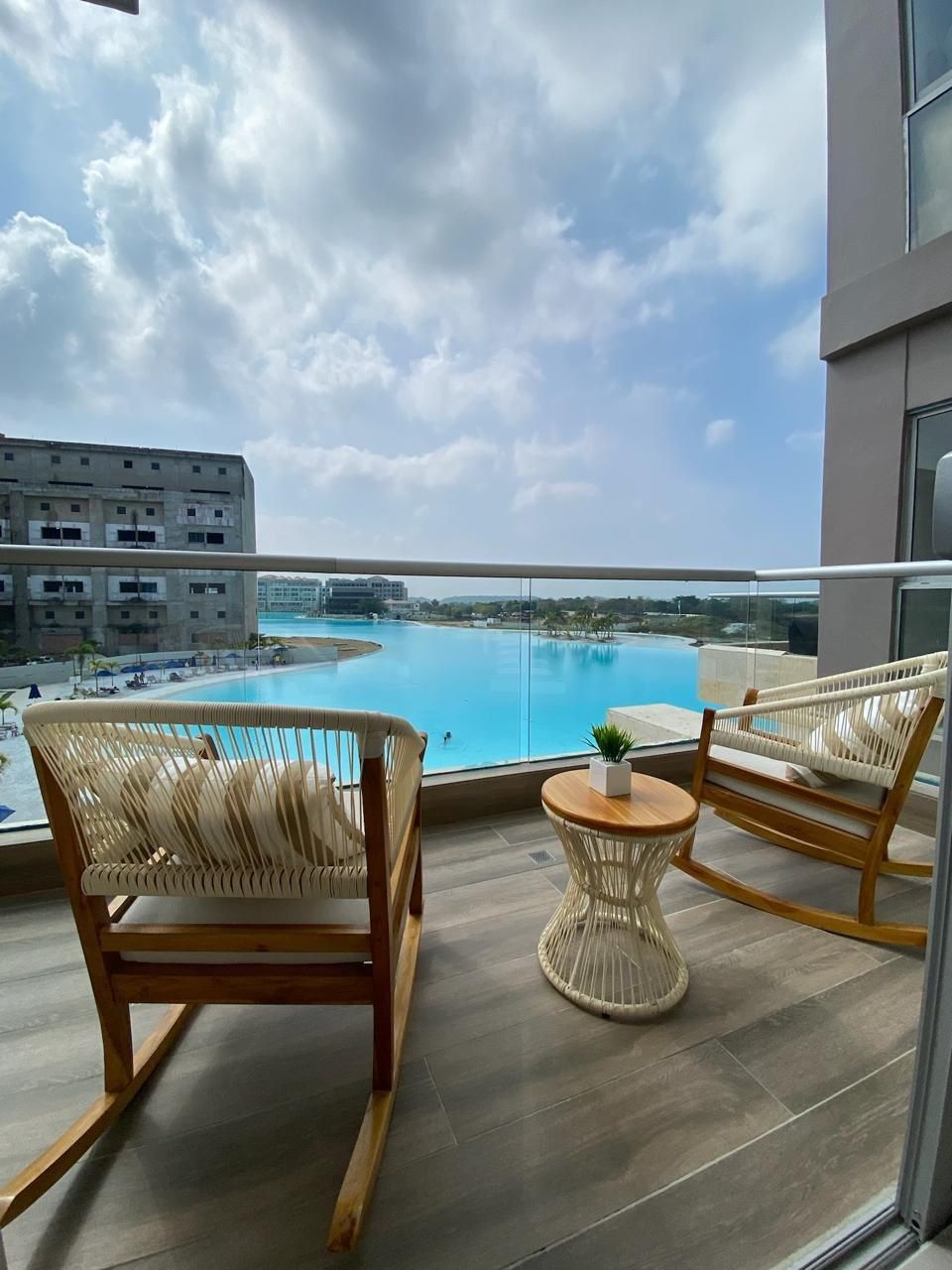 A balcony with wicker chairs and a small round table, overlooking a swimming pool and a distant cityscape.