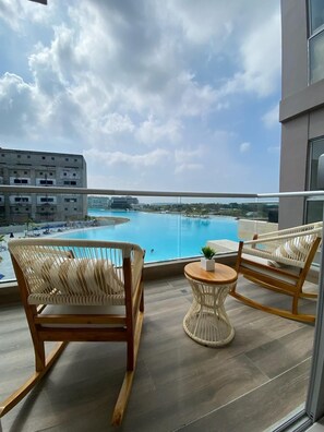 Un balcon agrémenté de fauteuils en osier et d’une petite table ronde, offrant une vue sur une piscine et sur le paysage urbain qui s’étend au loin.