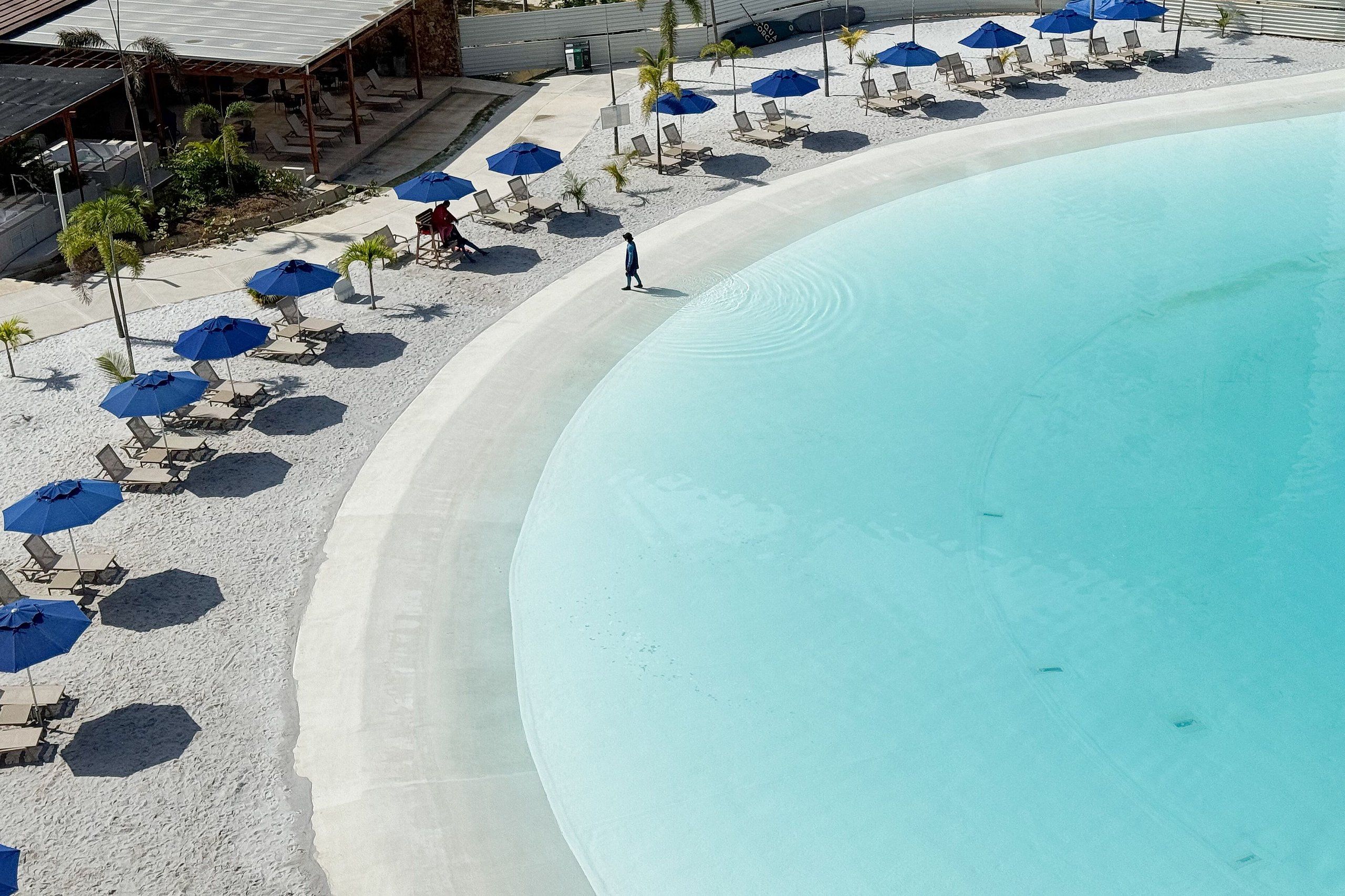 A poolside area with lounge chairs and umbrellas, a sandy beach, and a clear blue pool.
