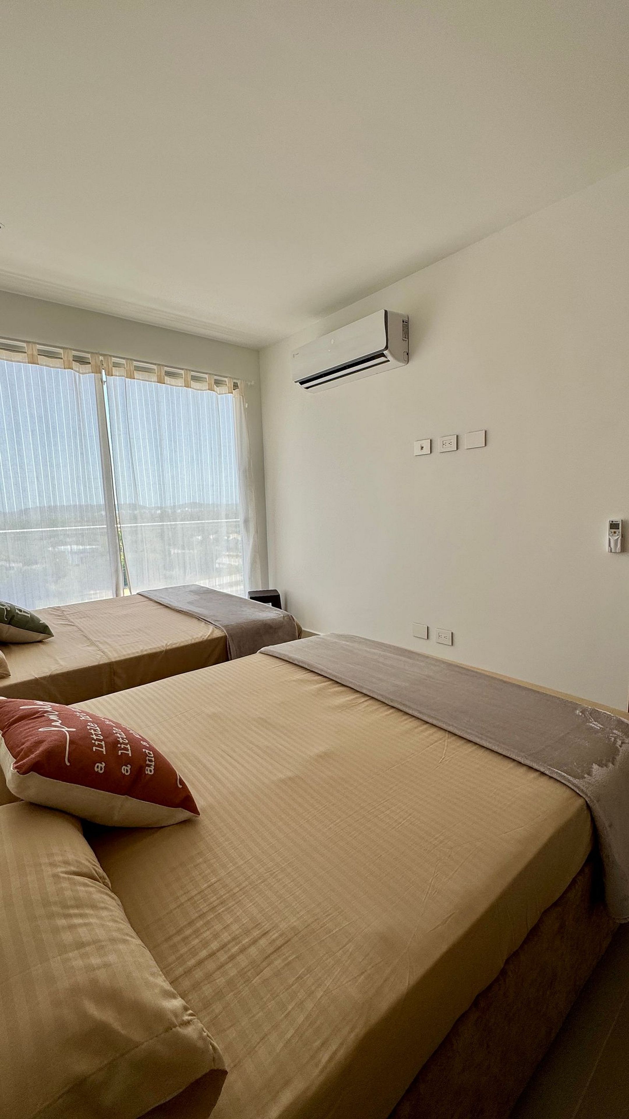 A room with two beds, a window with curtains, an air conditioning unit, and a wall-mounted electrical outlet.
