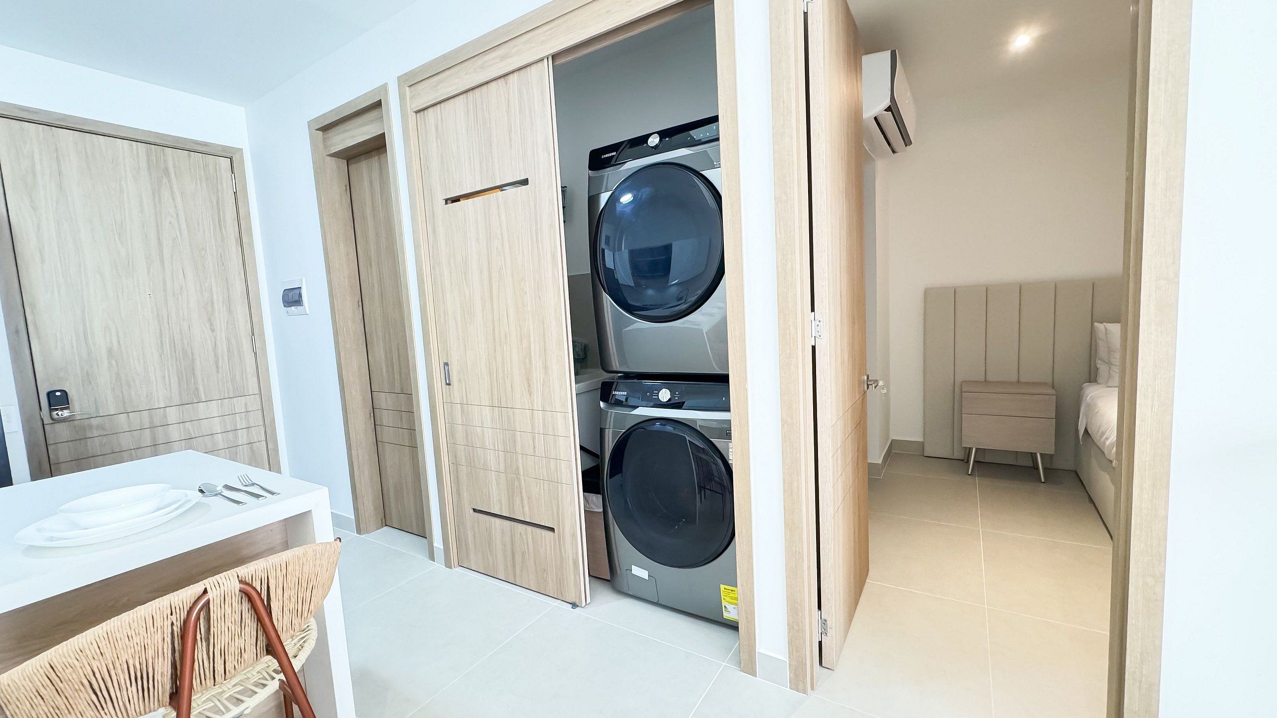 A compact laundry room with two front-loading washing machines, a wooden wardrobe, and a small bedside table with a lamp.