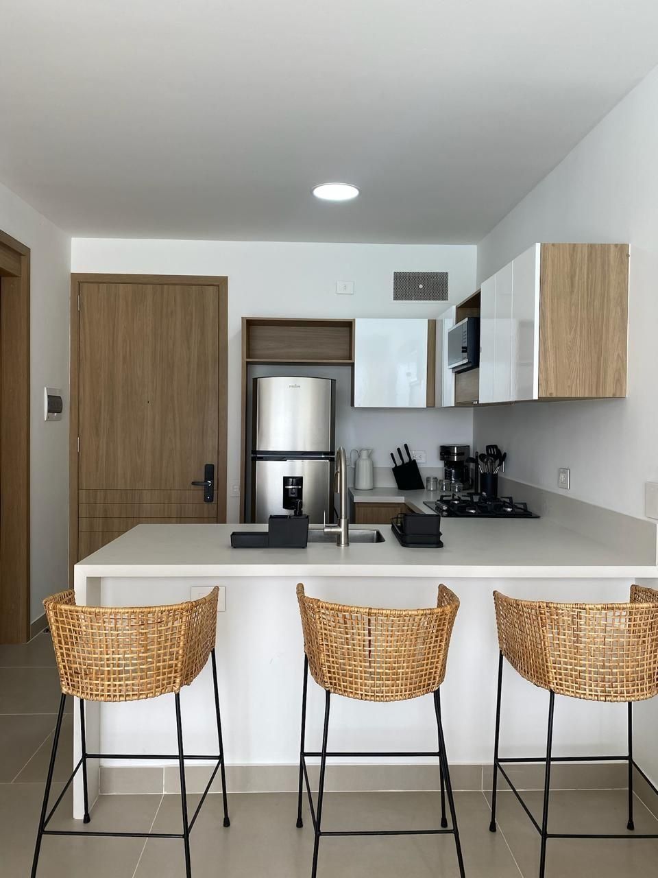A modern kitchen with a central island, stainless steel appliances, and three wicker bar stools.