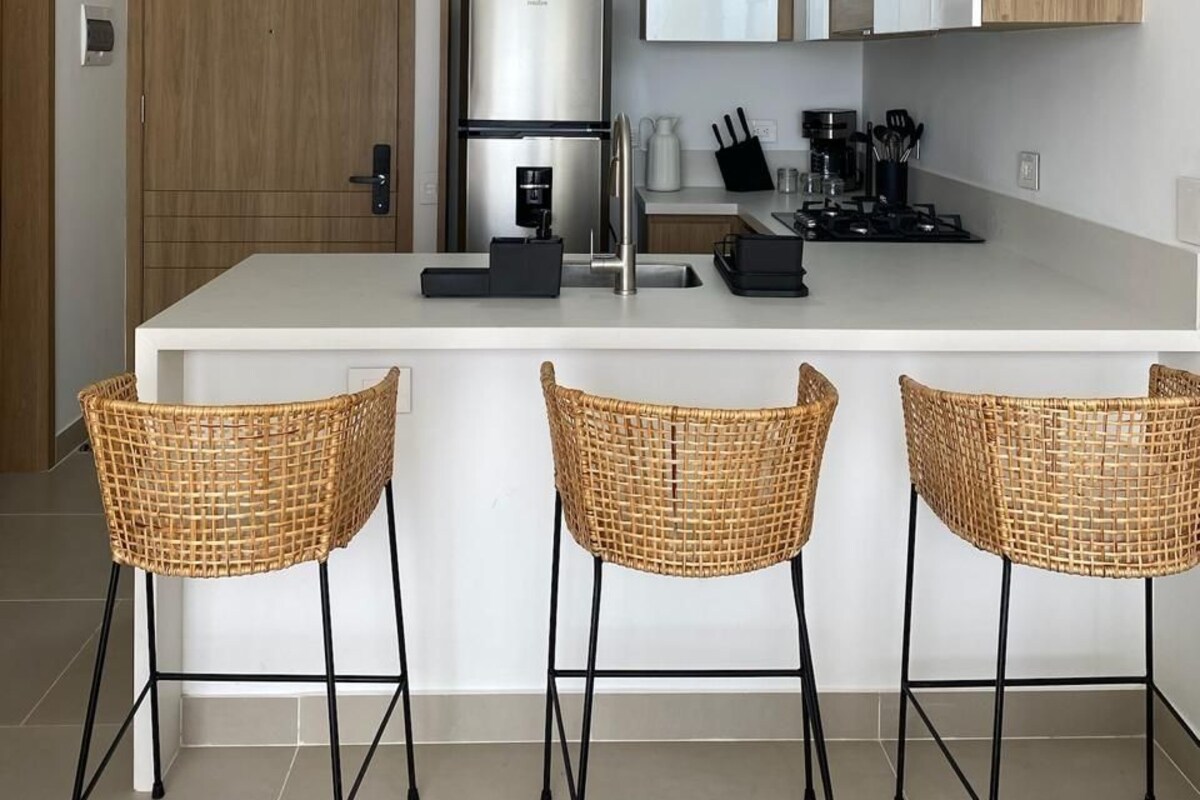 A modern kitchen with a central island, stainless steel appliances, and three wicker bar stools.