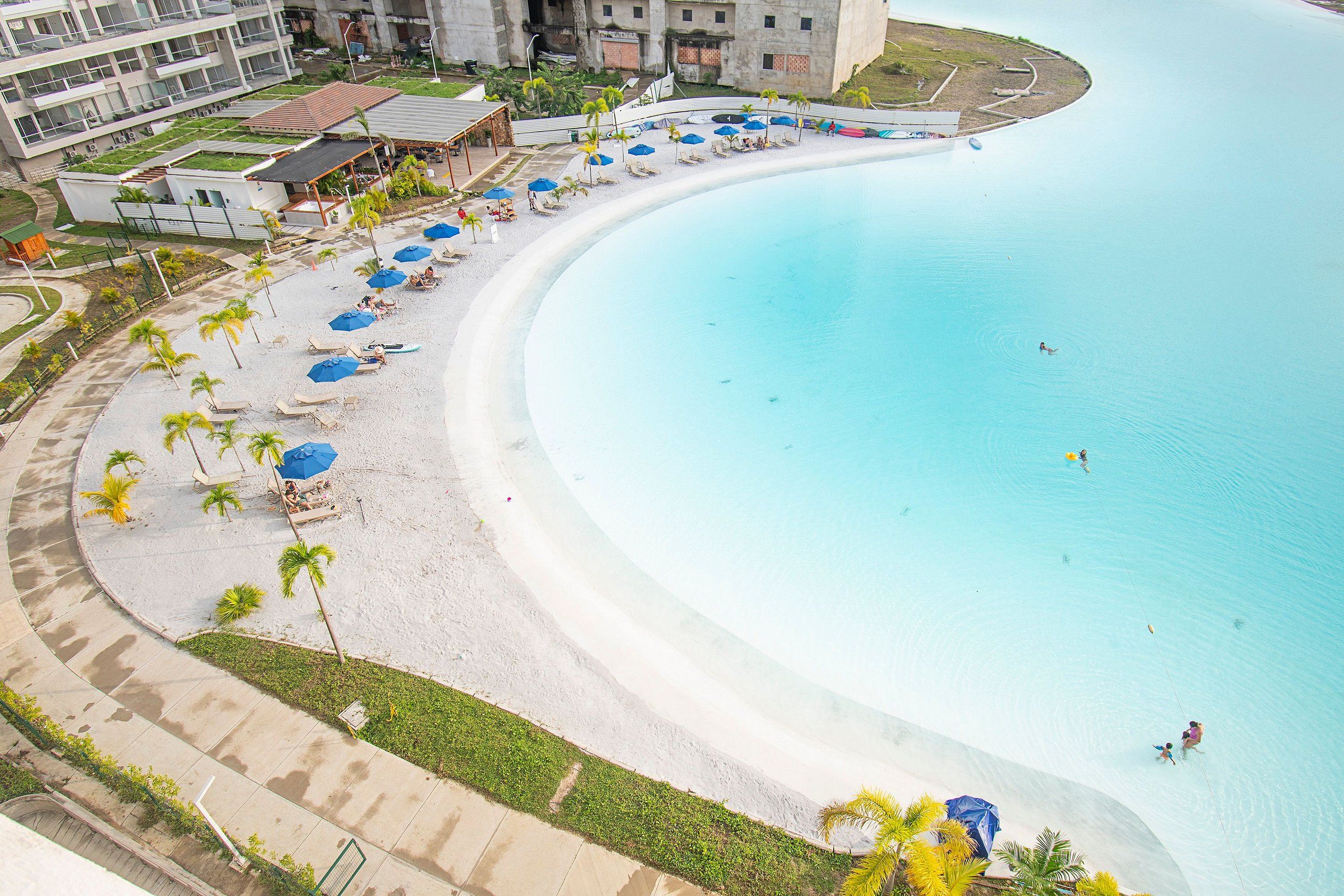 Une plage en forme de courbe, avec une piscine, des palmiers et des chaises de plage.