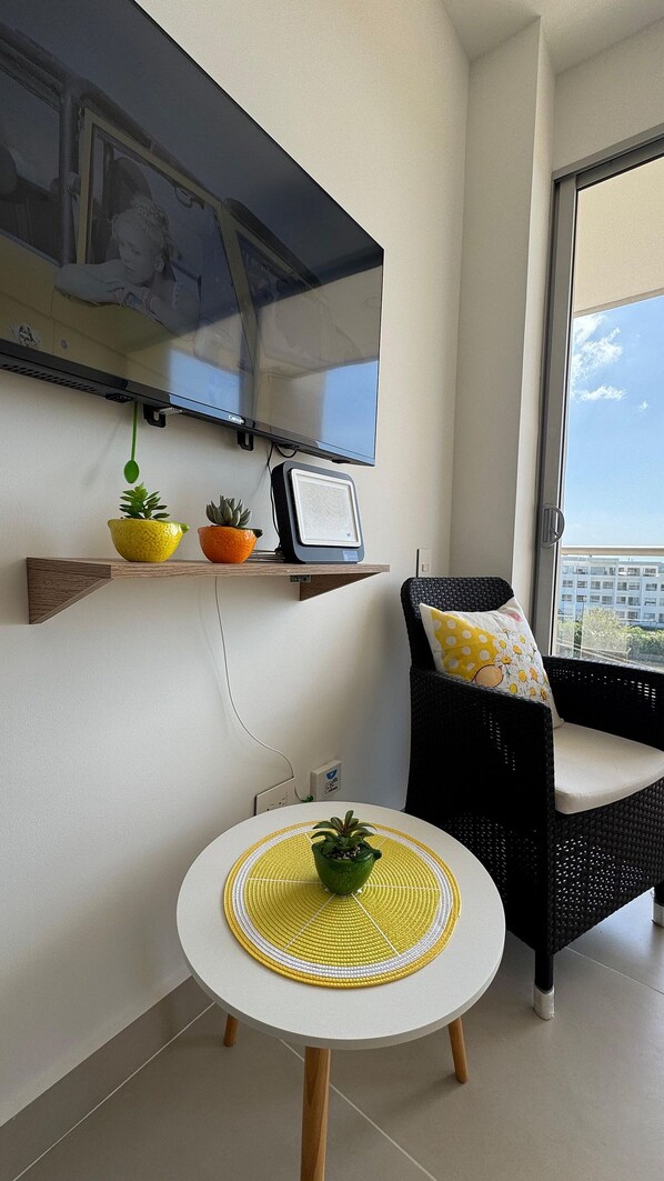 Un salon moderne avec un téléviseur à écran plat fixé au mur, une petite table surmontée d’un set de table en fibres tressées, un fauteuil en osier noir avec un coussin, et un balcon offrant une vue sur les immebles.