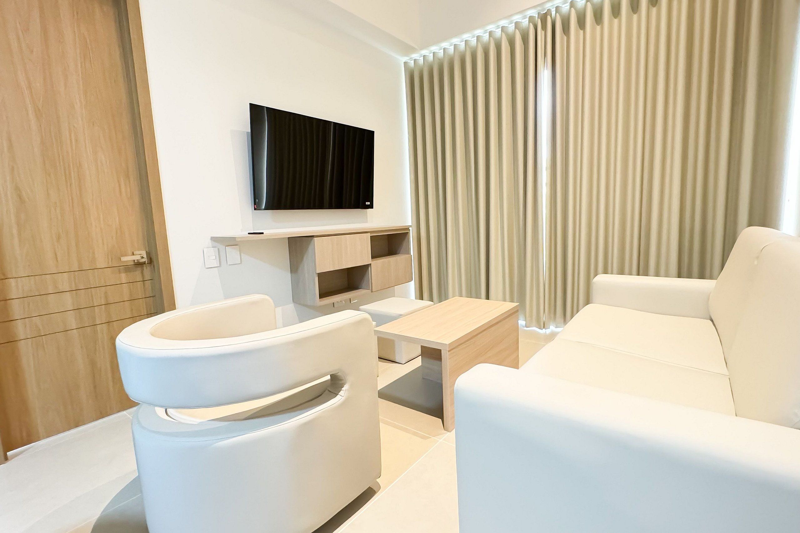A modern living room with a white sofa, a white armchair, a wooden coffee table, a flat-screen TV mounted on the wall, and a large window with vertical blinds.