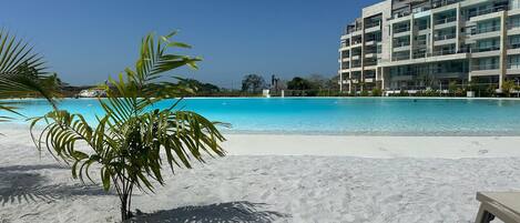 Una playa con una piscina azul cristalina, palmeras y un hotel de varios pisos al fondo.