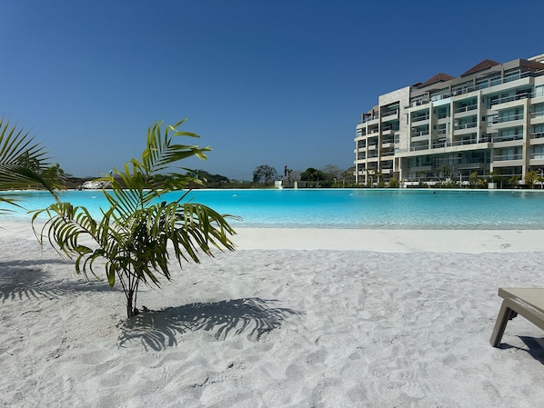 A beach with a clear blue pool, palm trees, and a multi-story hotel building in the background.