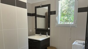 A bathroom with a white sink, dark cabinetry, a wooden trash bin, and a window with a view of greenery.