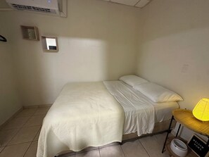 A small, clean room with a single bed, a bedside table with a lamp, and a small shelf.