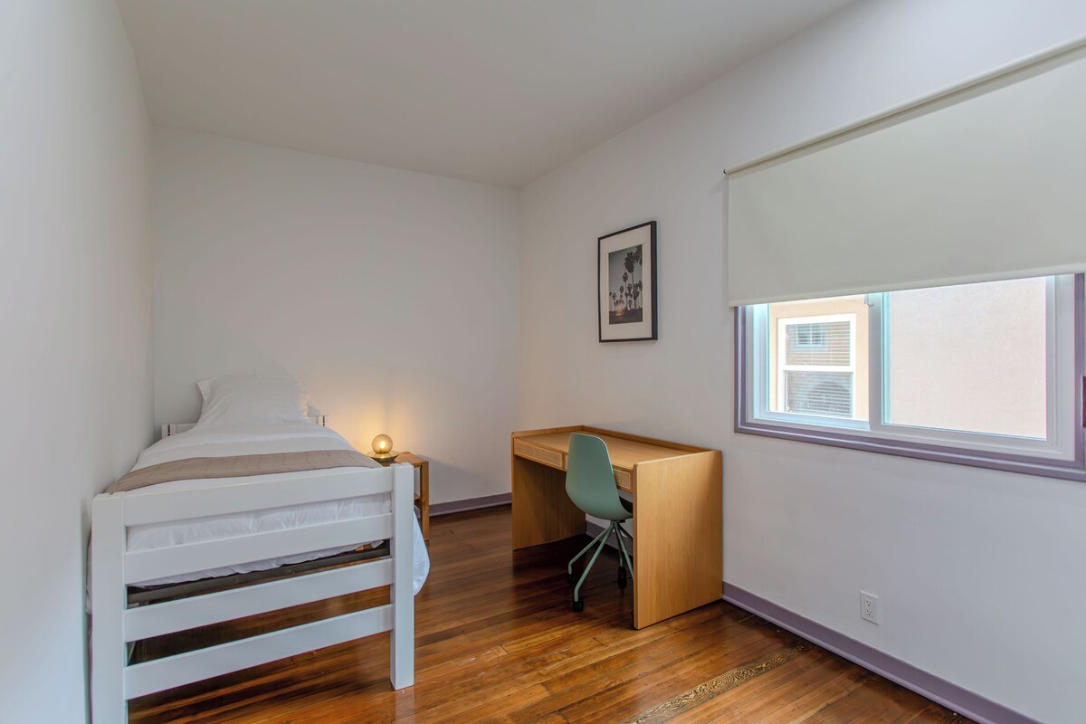 A bedroom with a bunk bed, a desk, and a chair. There is a window with blinds, a framed picture on the wall, and a light on the desk.