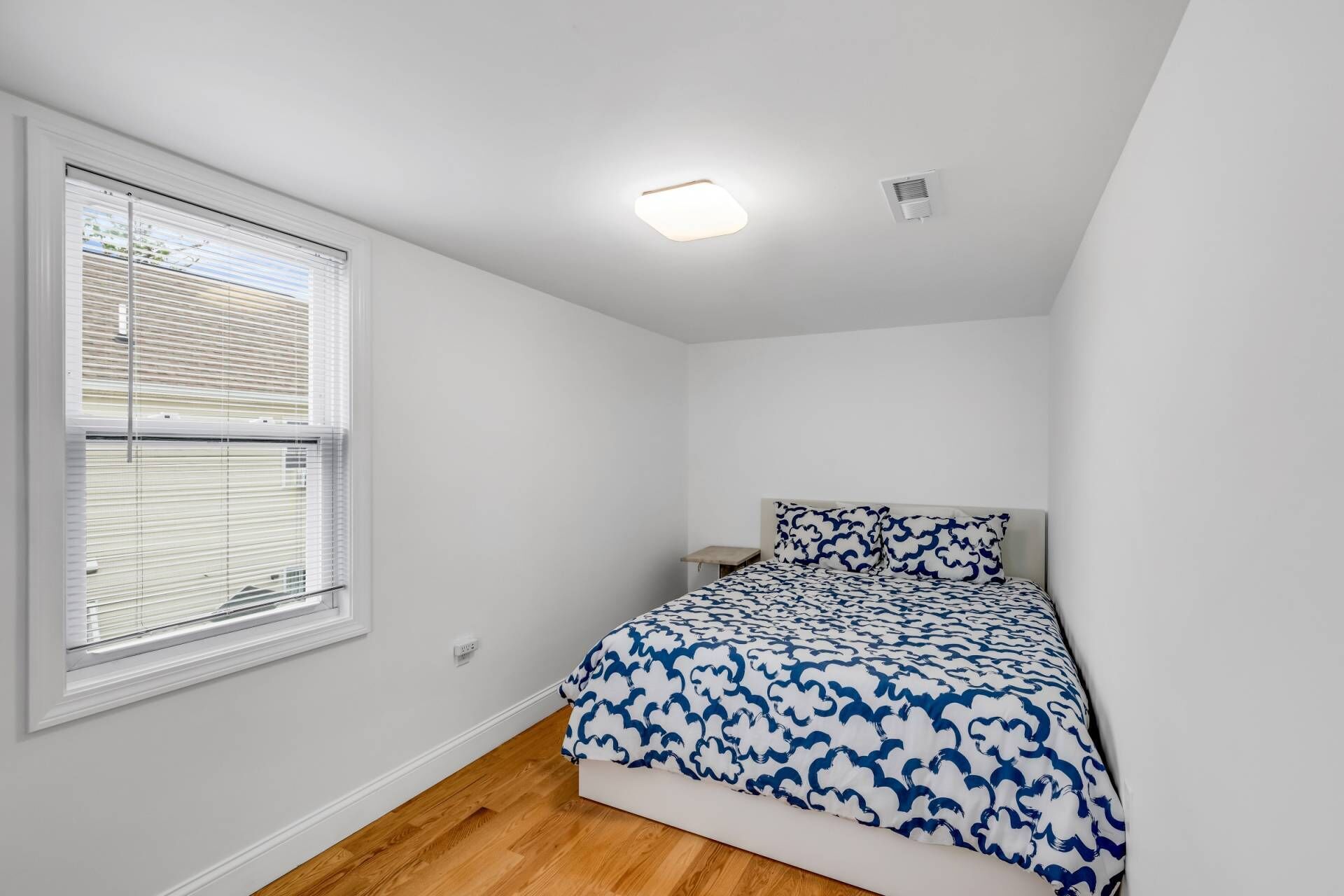 A small, white-walled bedroom with a single bed, a nightstand, and a window with blinds.