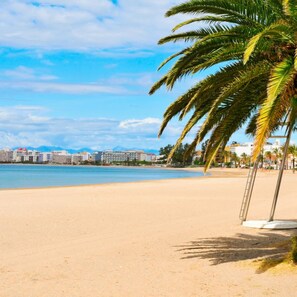 A beach with a palm tree, clear sky, and buildings in the background. - Norfeu Park 2-4 in Roses (Roses)