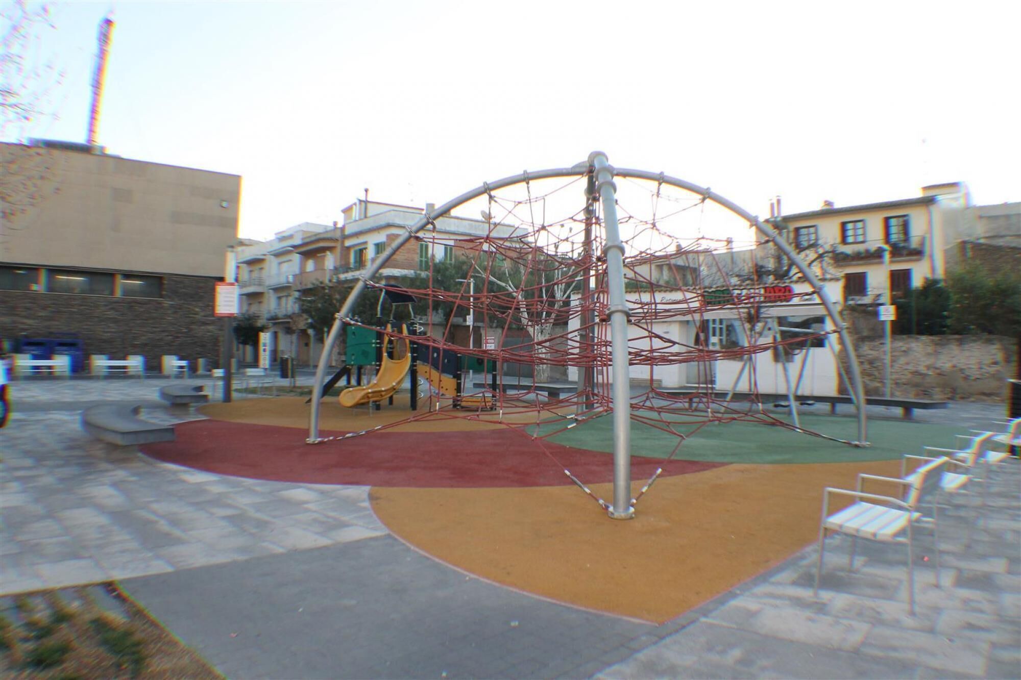 A playground with climbing structures and slides, surrounded by a paved area with benches.