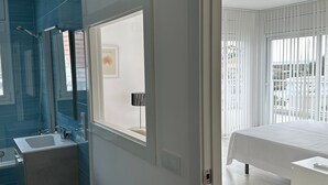 A modern bathroom with a shower, sink, and mirror. There is a door leading to a bedroom with a bed and a window. - Cap Llarg 9 in Roses (Roses)