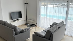 A modern living room with two grey sofas, a flat-screen TV, a floor lamp, and a view of a pool through large windows. - Cap Llarg 9 in Roses (Roses)
