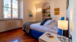 A bedroom with a large window, a radiator, a bed with blue bedding, a bedside table with a book, and wall-mounted shelves with plants and artwork. - Gerbett Apartment with Terrace by Wonderful Italy (Como)