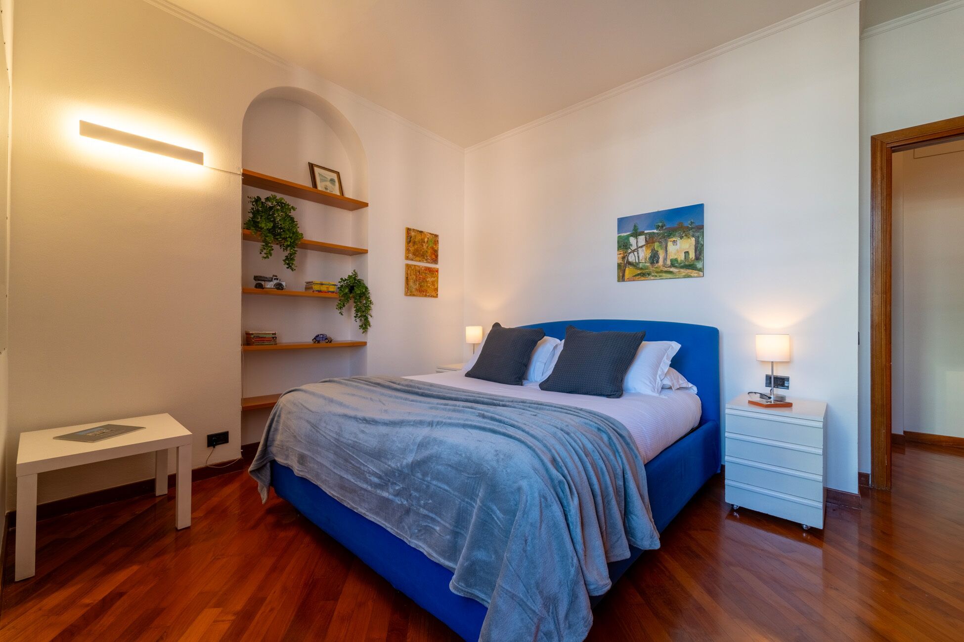A bedroom with a bed, bedside tables, a small white table, shelves with plants, and a painting on the wall.