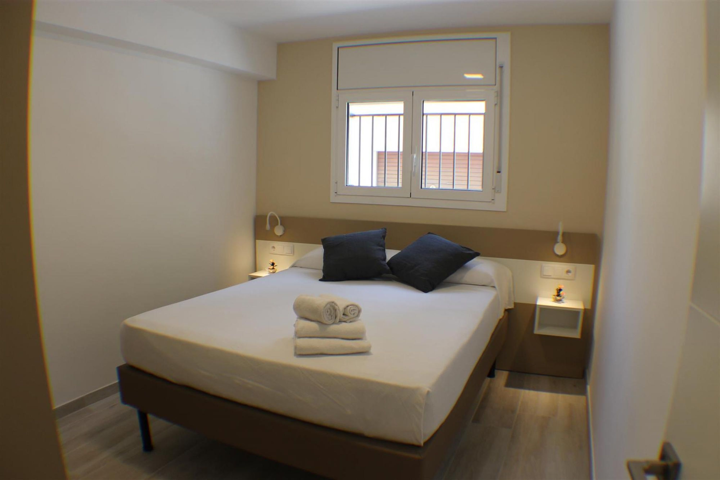 A neatly made bed with a white mattress and dark pillows, a small bedside table with a lamp, and a window with blinds.
