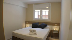 A neatly made bed with a white mattress and dark pillows, a small bedside table with a lamp, and a window with blinds. - Cosconilles Bajo in Roses (Roses)