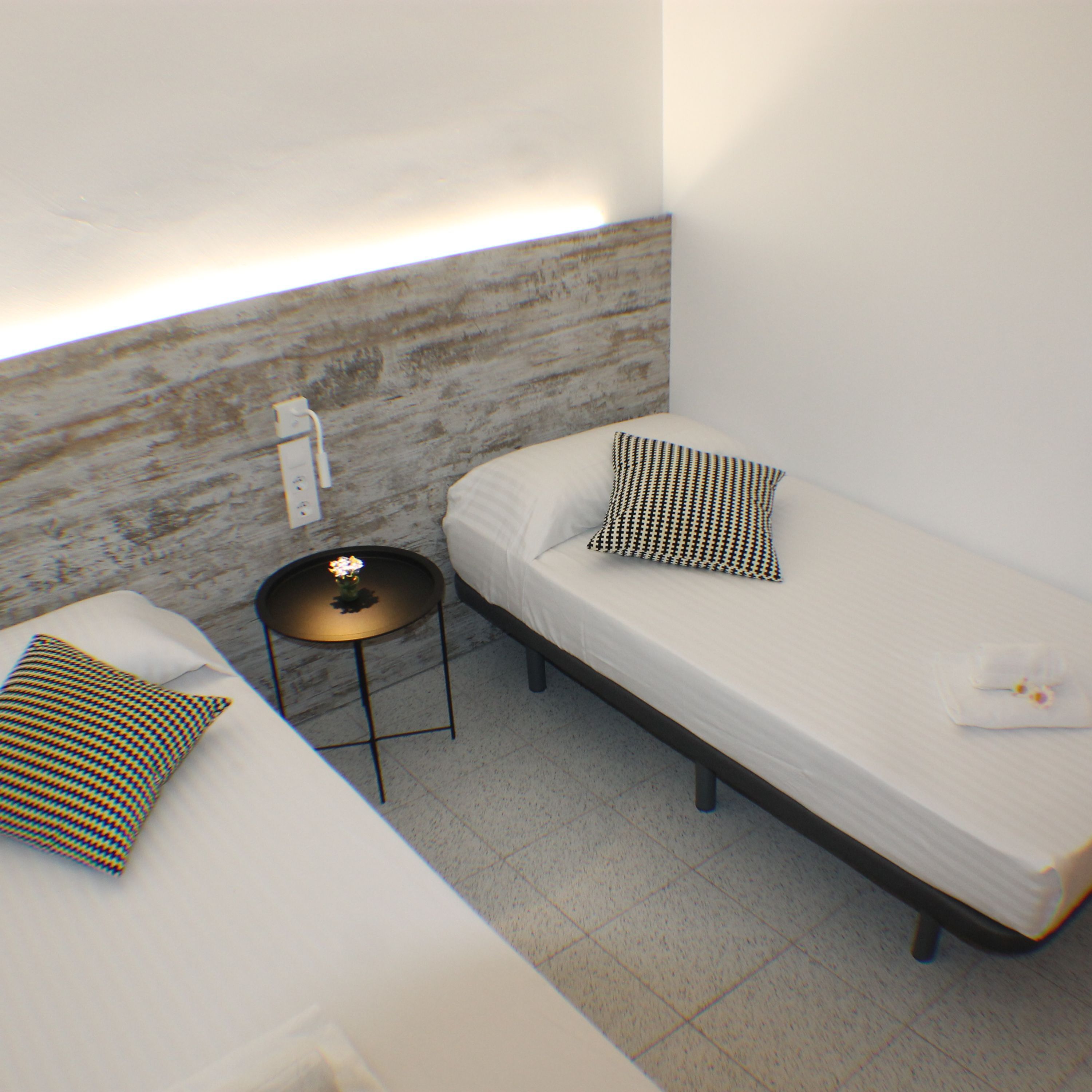A small, modern room with two single beds, a small round table with a flower, and a wall-mounted light fixture.