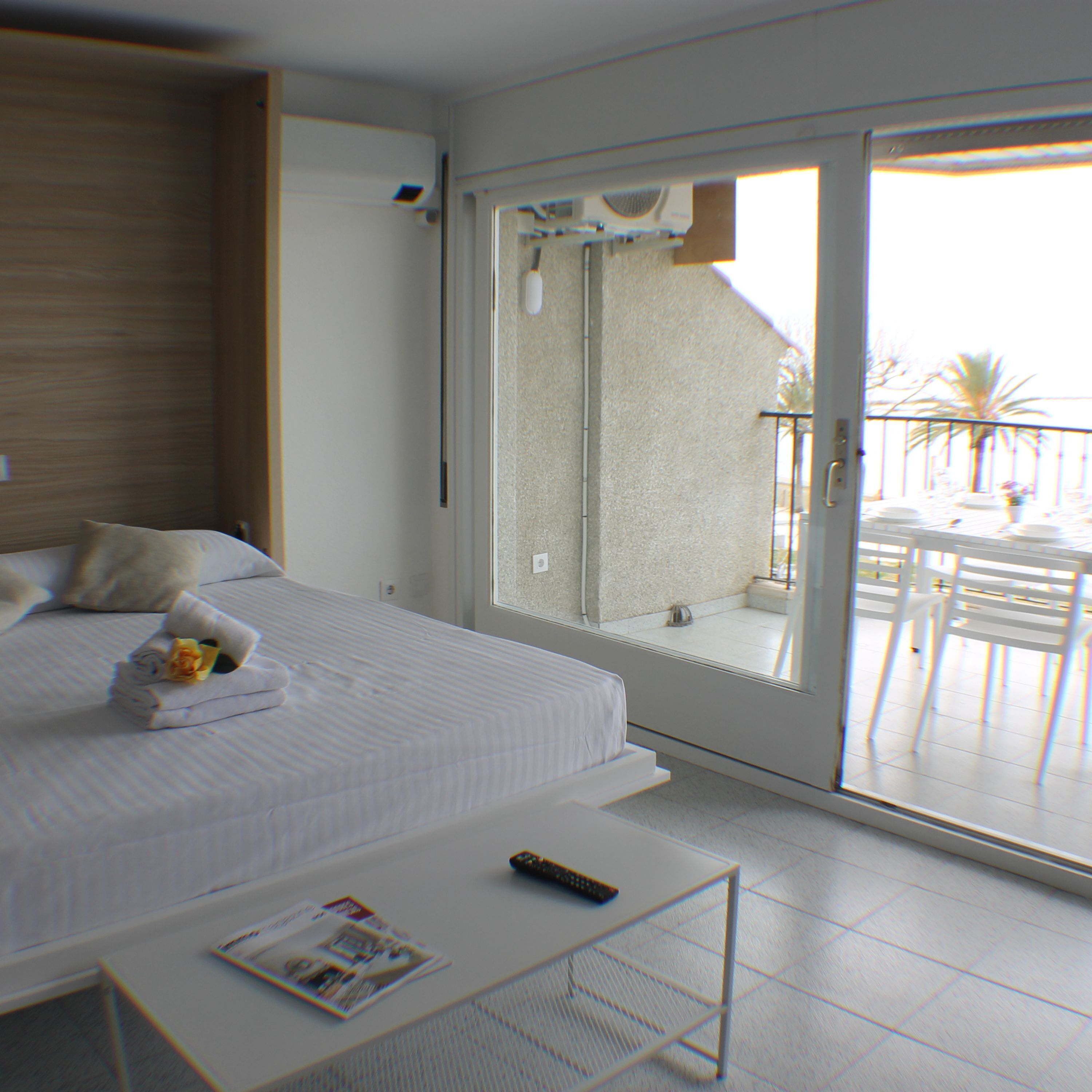 A bedroom with a bed, a bedside table, a remote control, and a window leading to an outdoor balcony with chairs and a table.