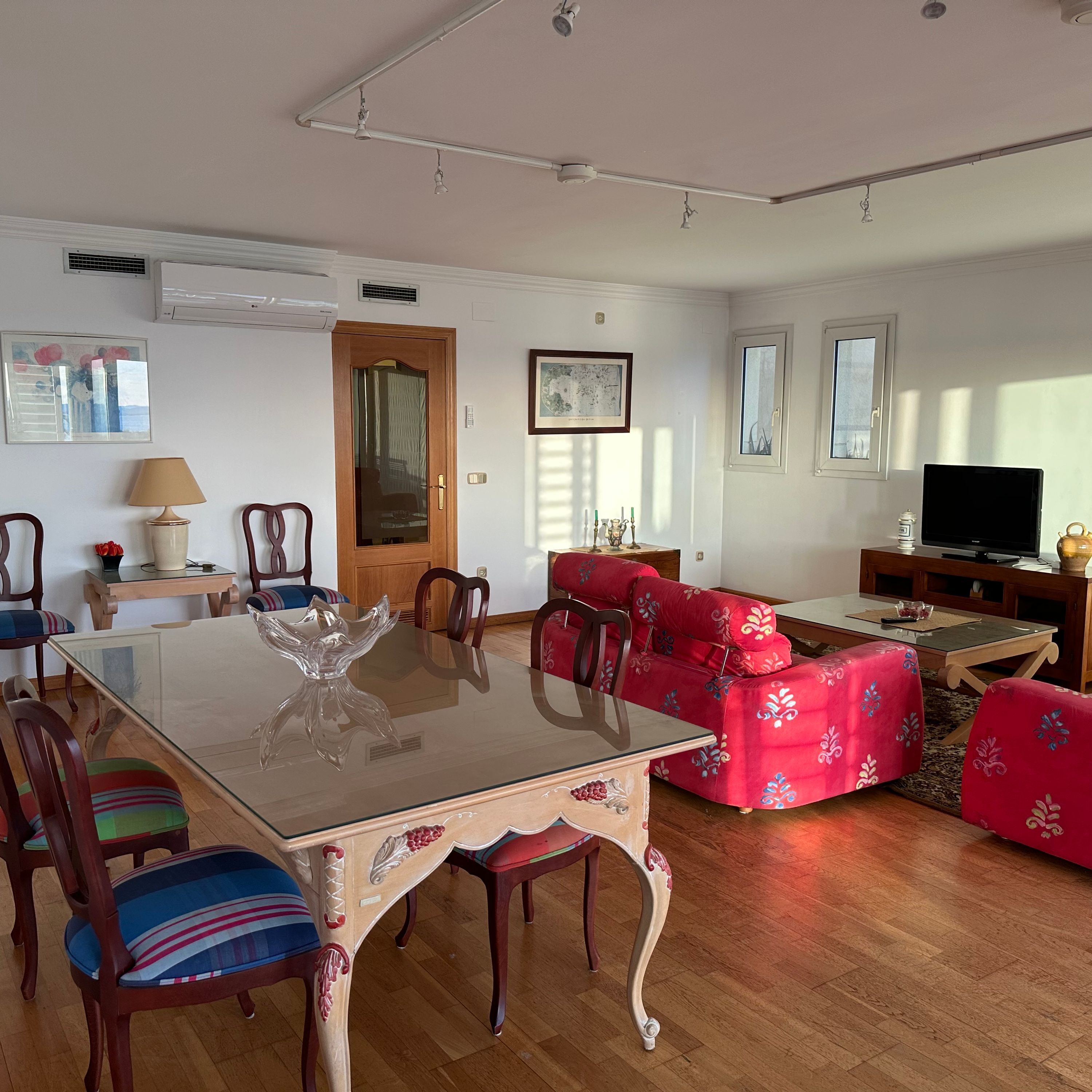 Uma sala de estar ampla com uma mesa de jantar de vidro, sofá vermelho estampado e uma televisão sobre um móvel de madeira.
