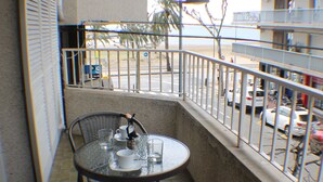Un balcon avec une table ronde et deux chaises en osier, donnant sur une rue bordée de voitures garées et agrémentée d’un palmier.