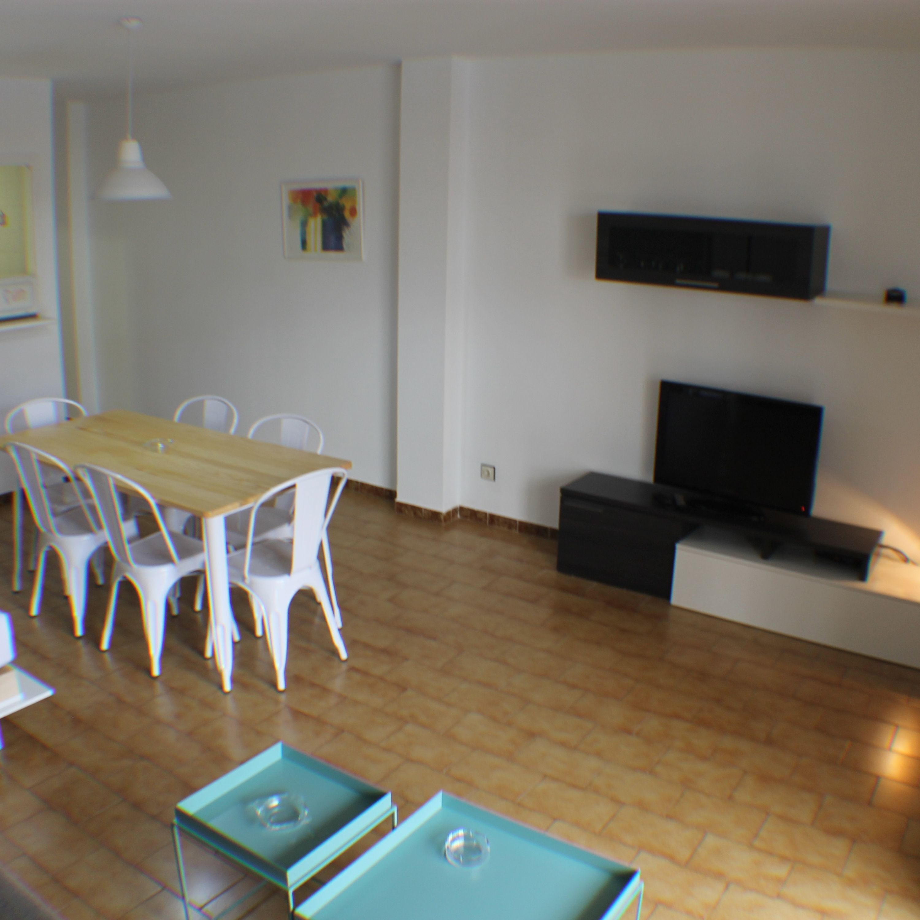 A dining area with a wooden table and white chairs, a television on a stand, and a tiled floor.