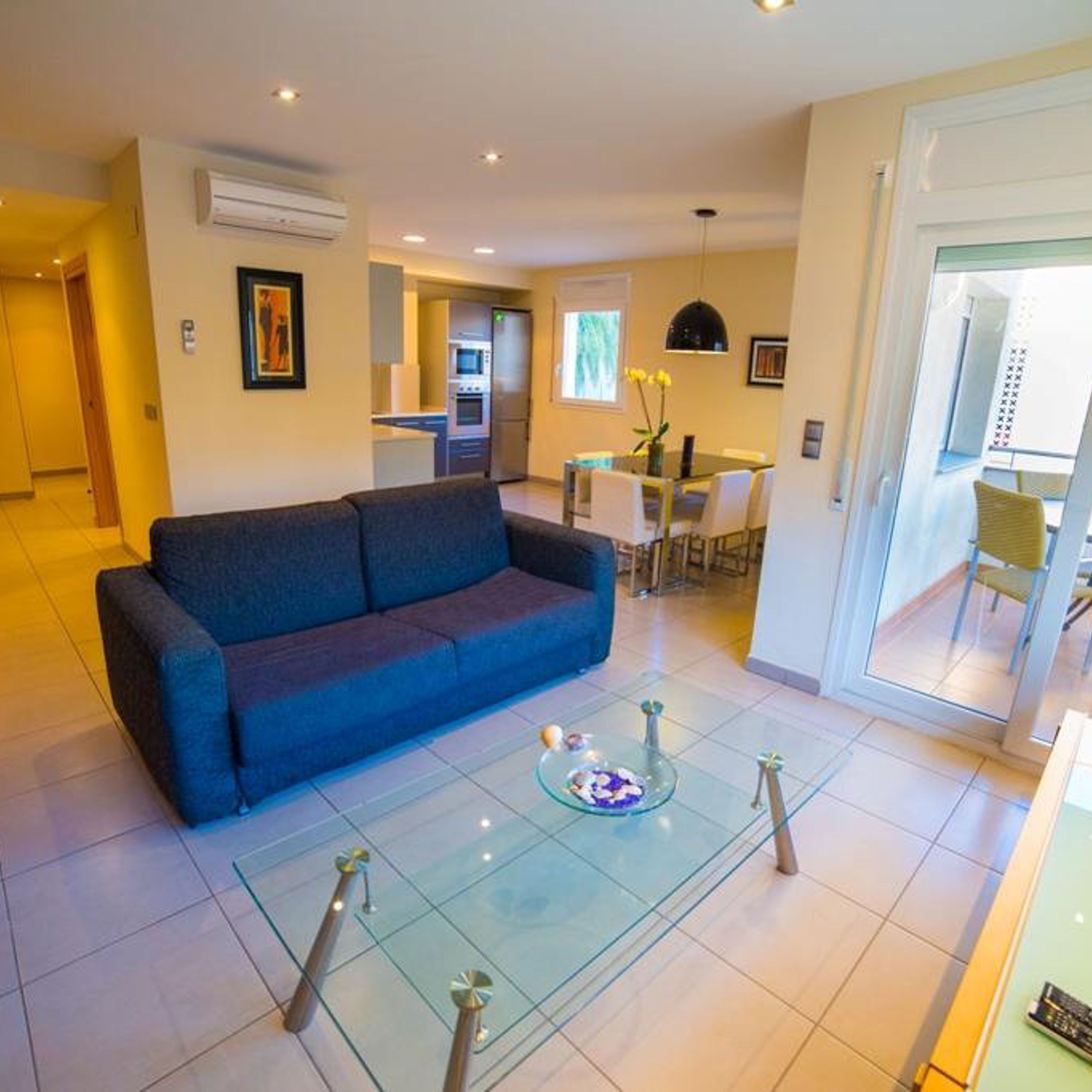 A modern living room with a blue sofa, a glass coffee table, and a dining area with a table and chairs.