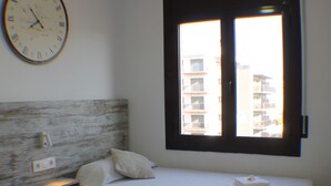 A hotel room with two single beds, a wooden headboard, a wall clock, and a window with a view of buildings.