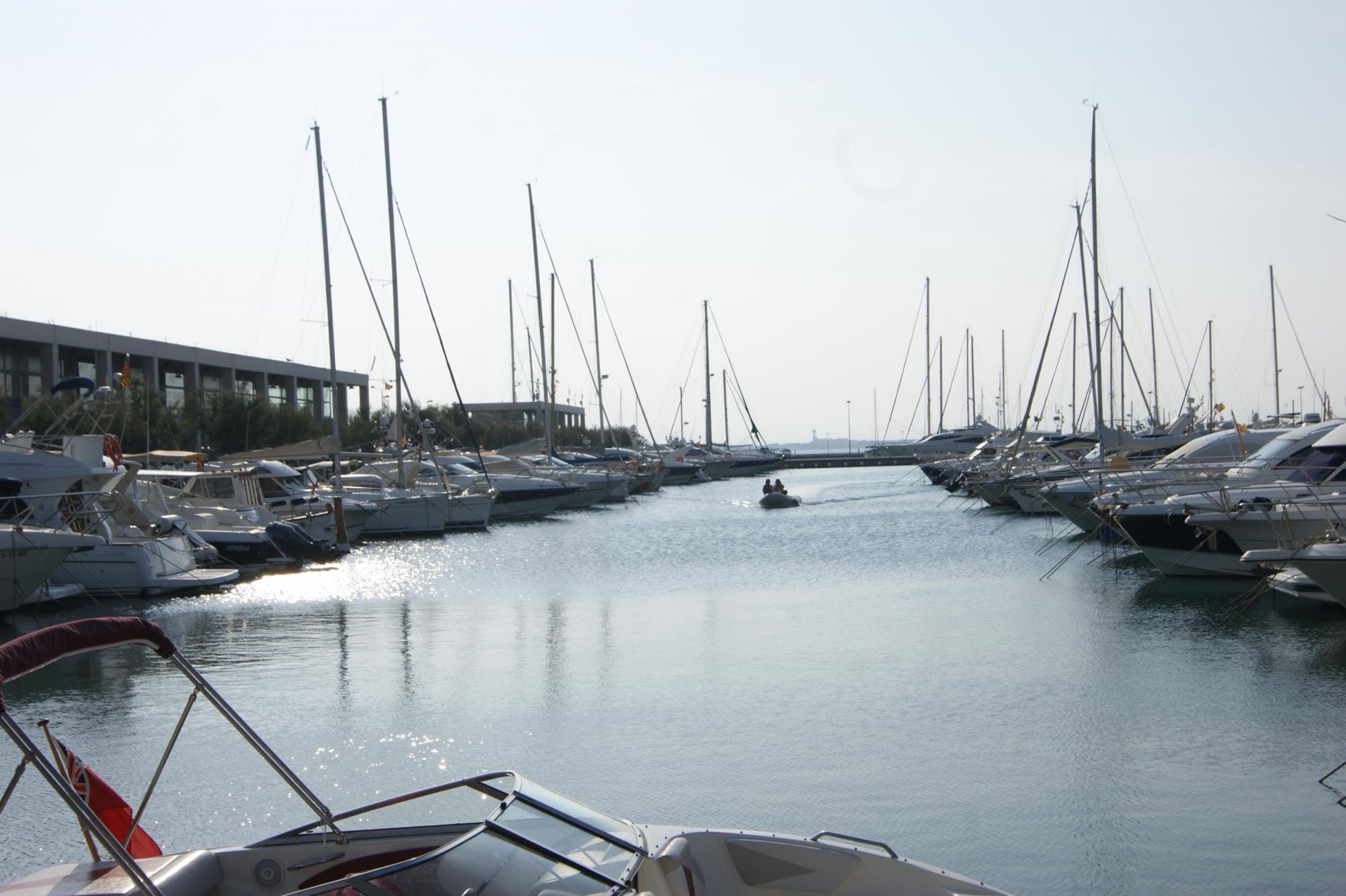 A marina with numerous boats docked in calm waters, a clear sky, and a building on the left.